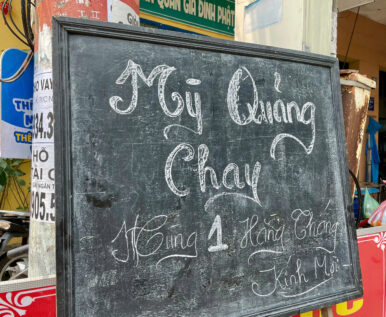 A close-up shot of a chalkboard menu board advertising "Mì Quảng Chay" (Vegetarian Quang Noodles) for the free monthly community charity kitchen